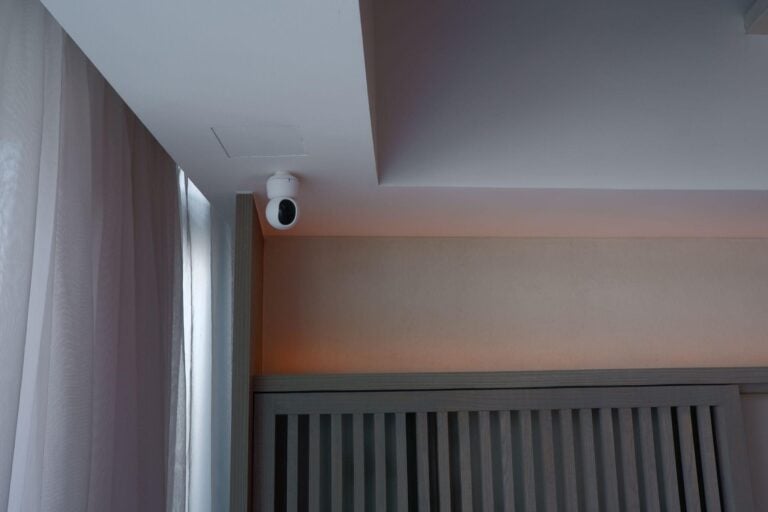 White indoor camera mounted upside down on the ceiling beside full-height curtains.