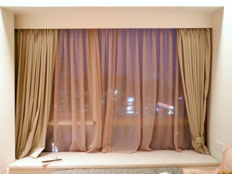 Bay window at night with sheer and blackout curtains drawn back and city lights outside.