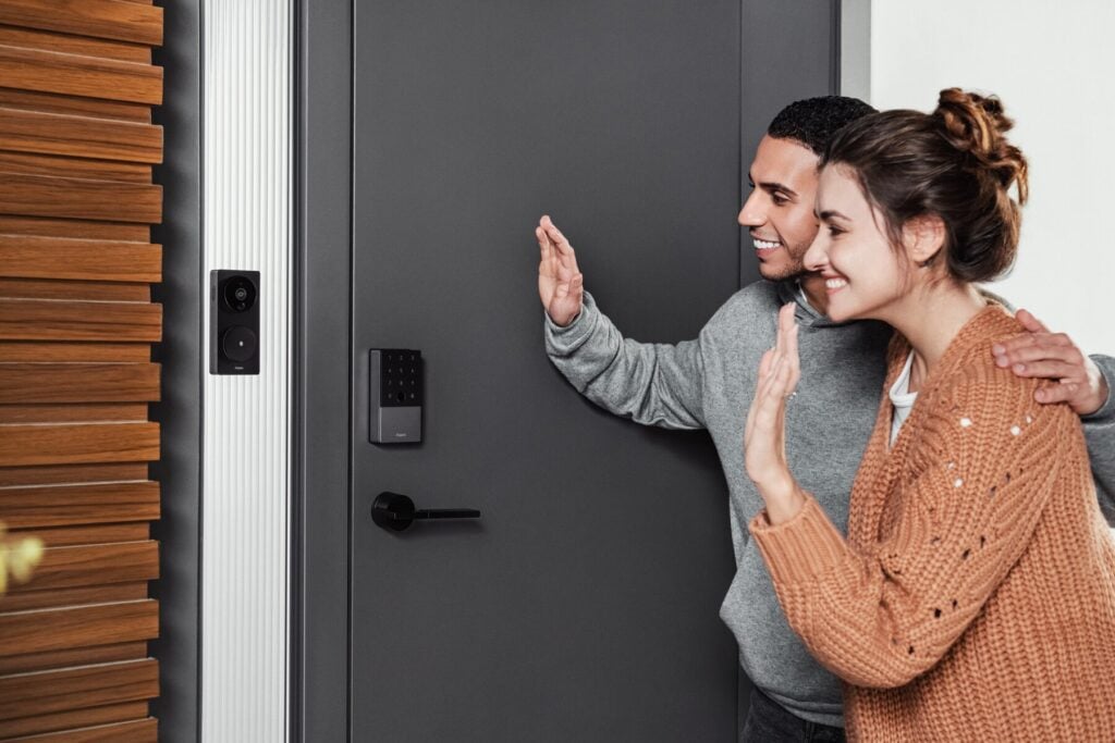 A couple waves at an Aqara video doorbell beside an Aqara keypad smart lock