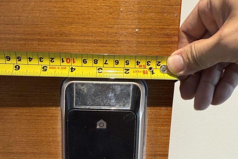 A hand measuring a door and its existing lock with a tape measure