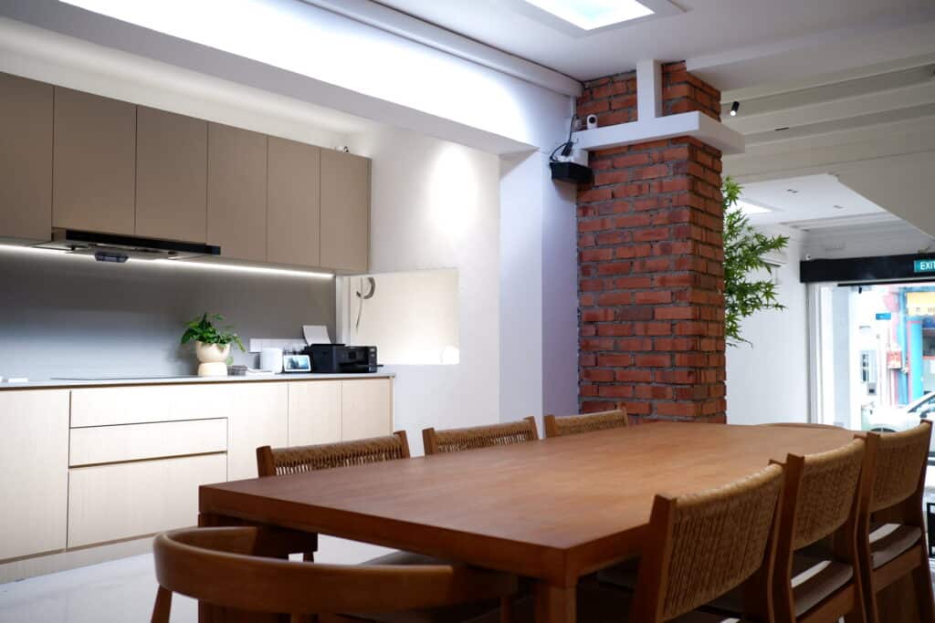 The dining area at the HomeSmart showroom, 24 Keong Saik Road: a long wooden table with woven chairs beside a kitchen counter and a brick column